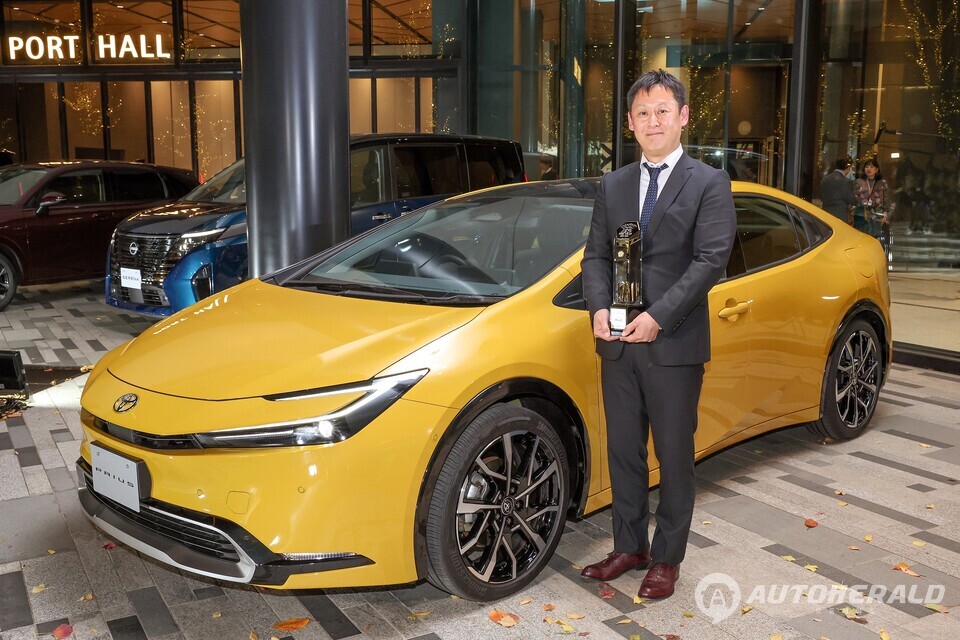 오야 사토기 프리우스 수석 엔지니어가 2023-2024 일본 올해의 차로 선정된 신형 프리우스와 함께 트로피를 들고 있다.